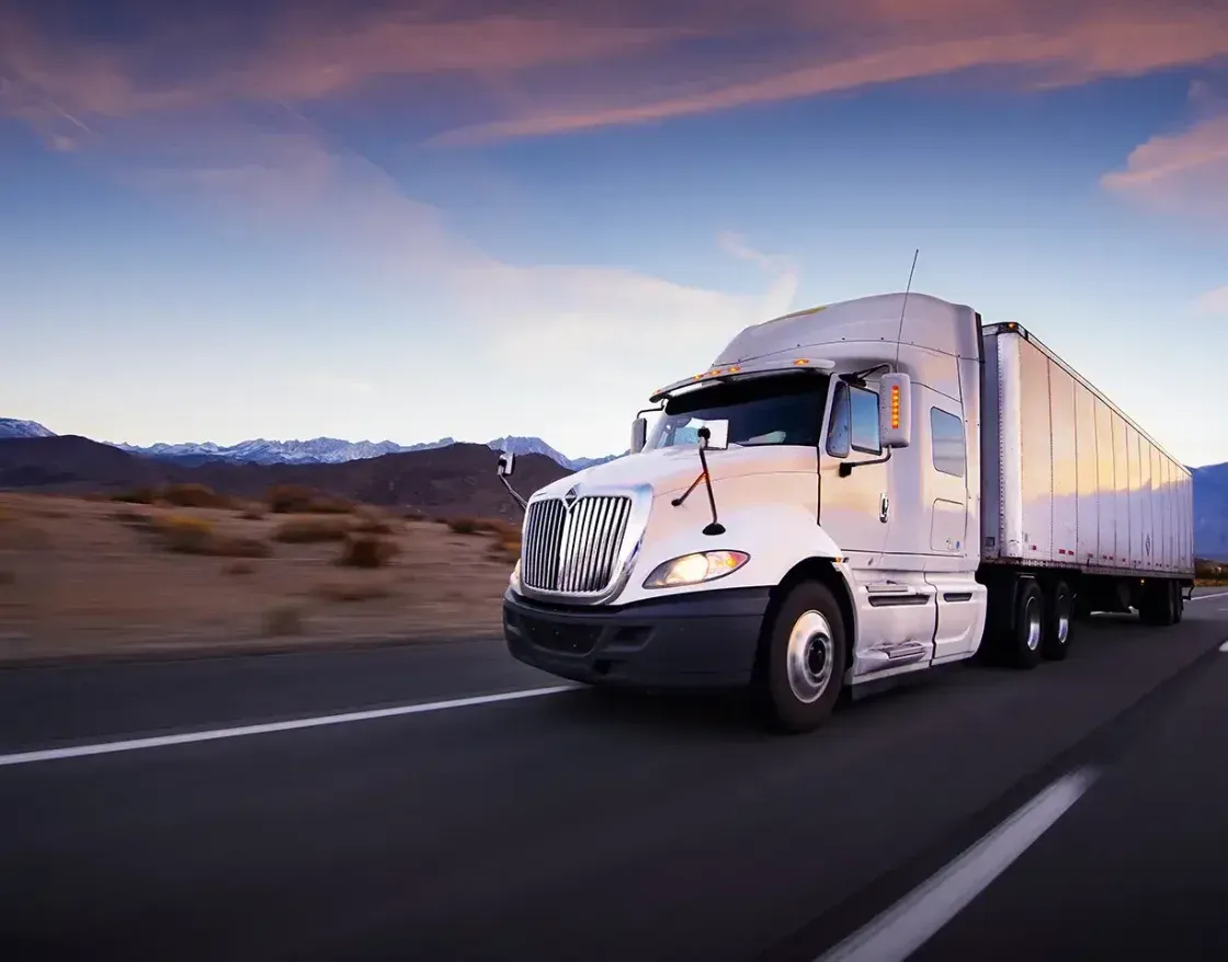 Semi truck pulling a white trailer down a desert highway at dusk